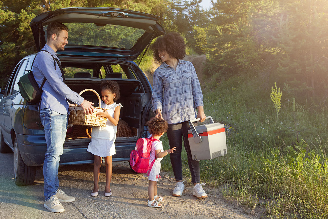 A family with two young children unpack coolers and snacks from the back of their car.