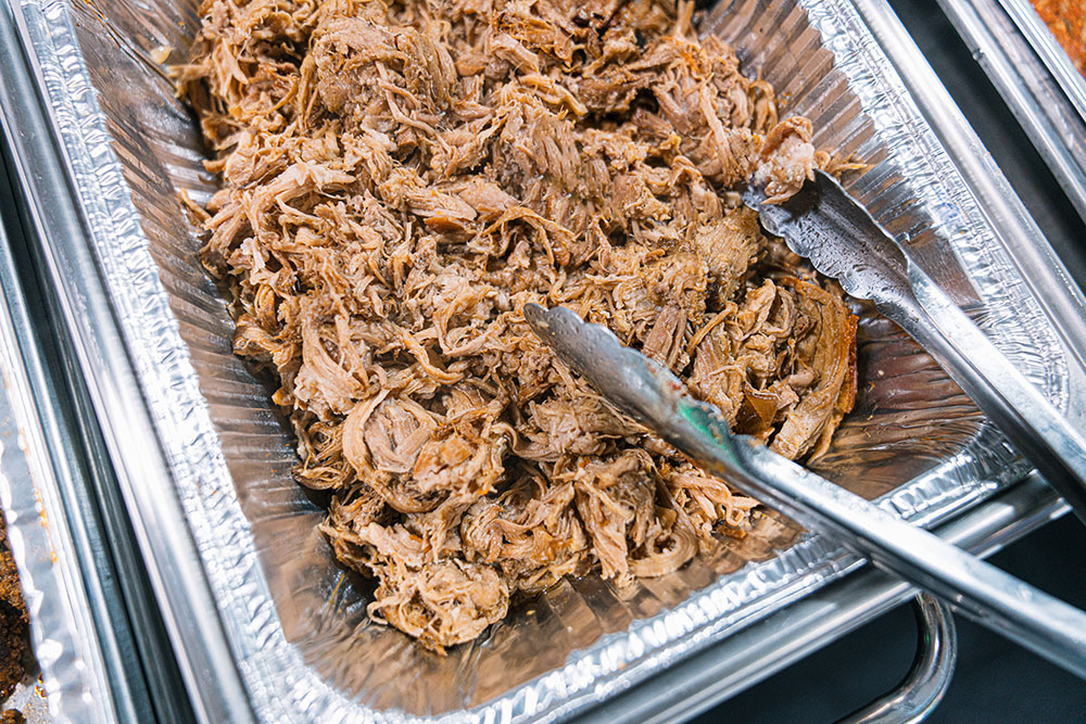 A buffet with pulled pork in an aluminum pan with tongs on the side.