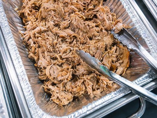A buffet with pulled pork in an aluminum pan with tongs on the side.